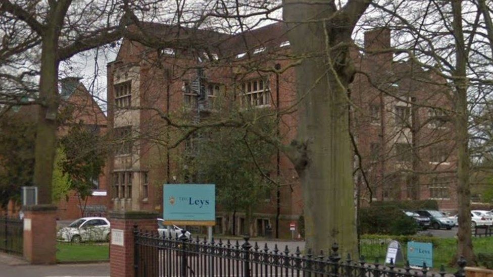 An exterior shot of The Leys school in Cambridge taken from the road. It shows a large brick building in the background with trees in the foreground and a blue school sign.