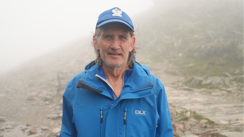 Volunteer national park warden Tony Ellis standing on the summit of Snowdon in heavy cloud, wearing a blue waterproof jacket and blue baseball cap