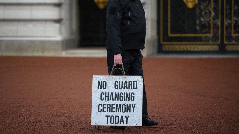 A police officer carries a sign saying 'no guard changing today' outside Buckingham Palace in London