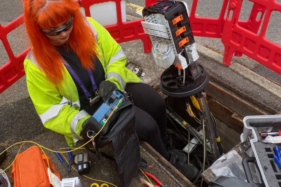 A female Openreach engineer wearing a high-vis vest examines a device in her lap tat is plugged into equipment coming out of the road where a steel plate has been removed for access
