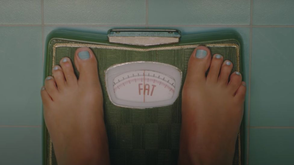 Скриншот музыкального клипа, показывающий напольные весы с надписью FAT