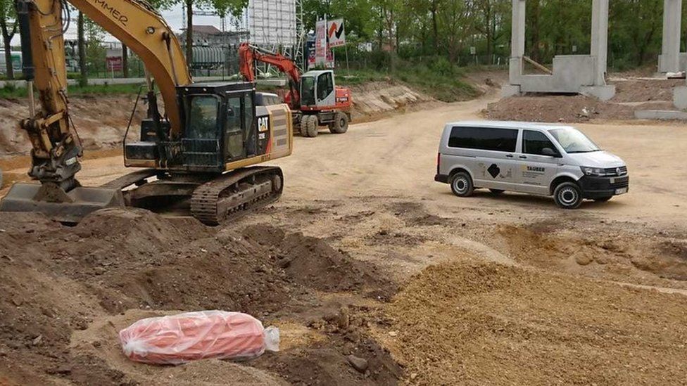 A consturction digger is seen, still and vacant, with its digging arm right next to an unexploded bomb wrapped in orange plastic