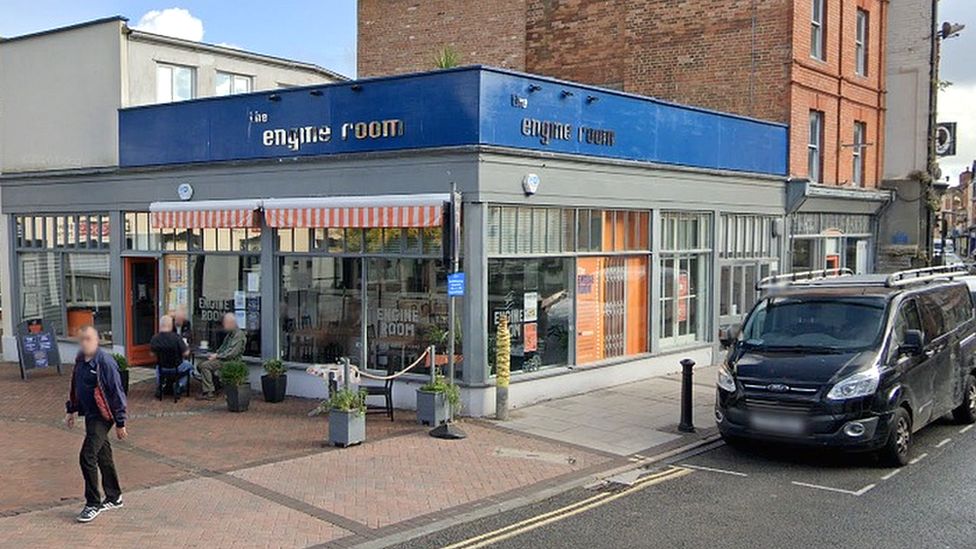 A single storey building with big window frontage on two sides, and blue backed signs above which say the engine room, some cafe tables and chairs outside the door