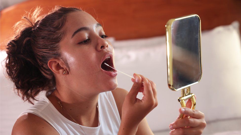 A woman swabs her throat while looking in a mirror