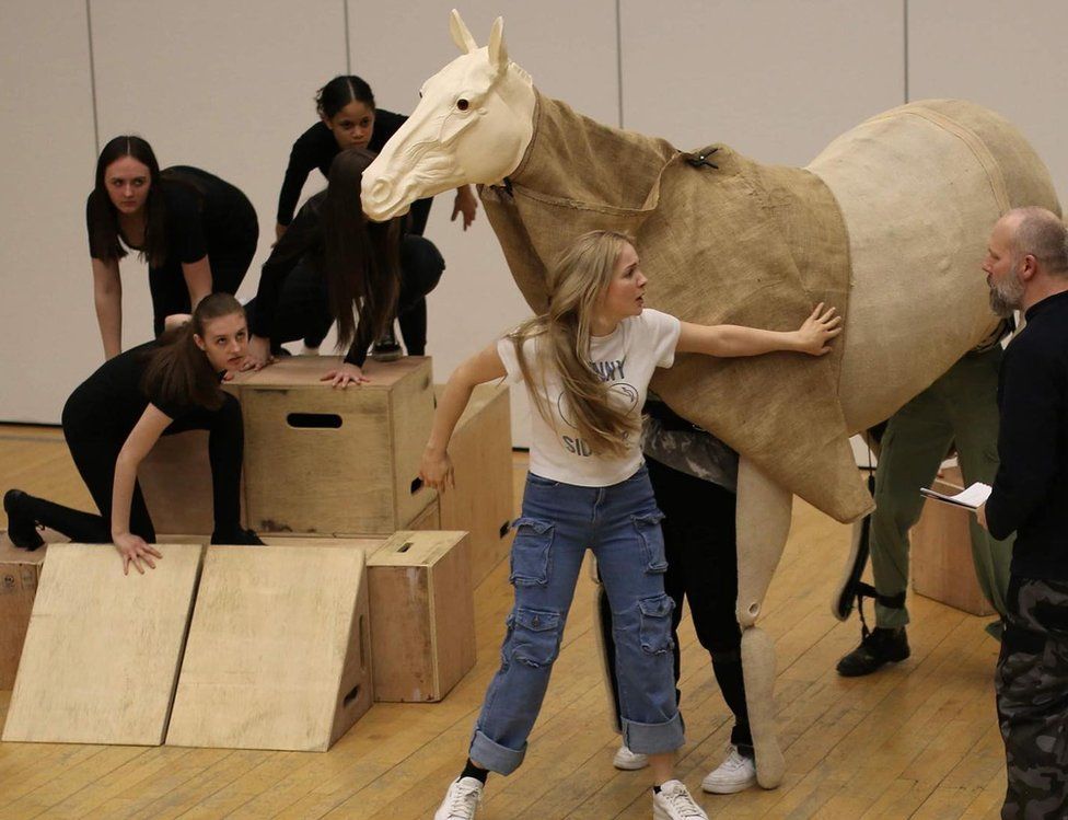 Actors dressed in black crouch on boxes next to a model of a white horse