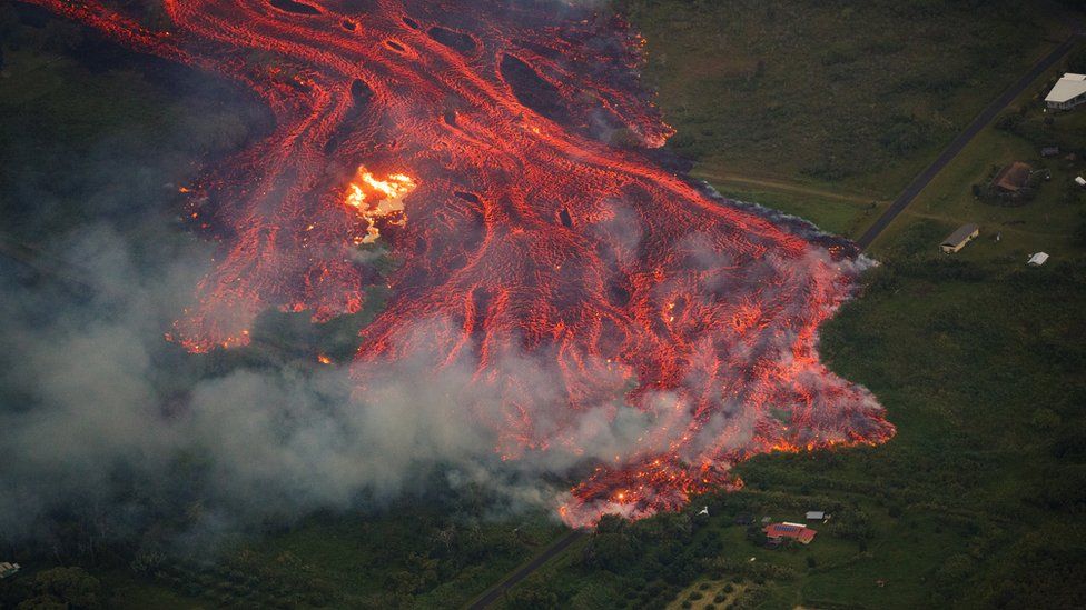 An aerial view of a massive flow of fast moving pahoehoe consuming everything in its path, as the flames from the remnants of one home burn