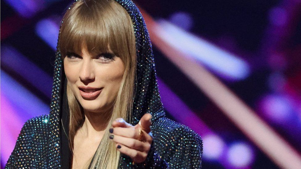 Taylor Swift points at the camera with a knowing smile. She's wearing a sparkly hooded dress with the hood pulled up