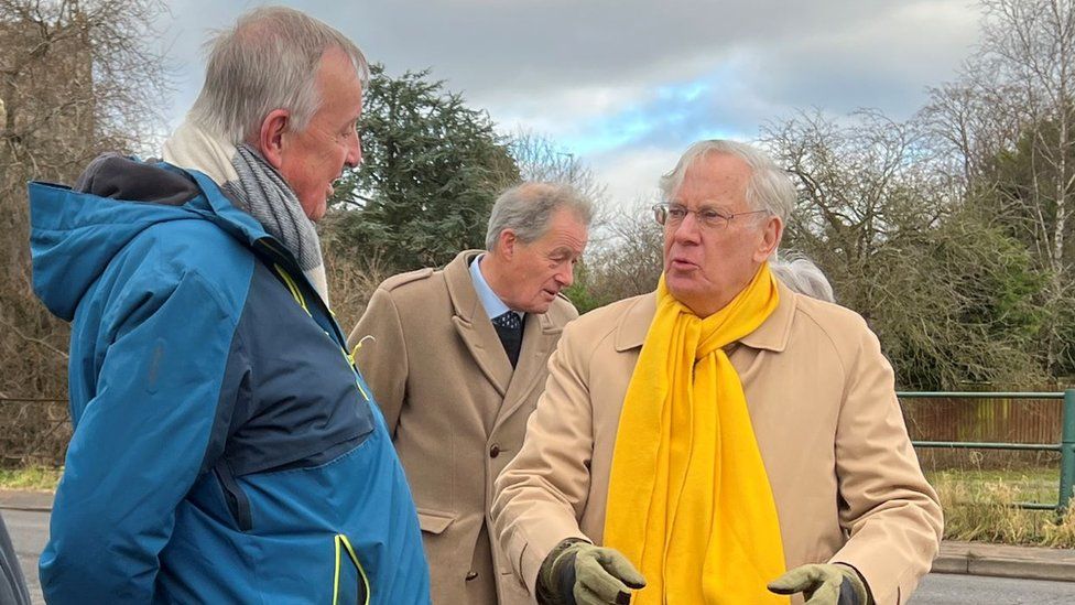 John Badham in a blue coat speaking with the duke, who wears a yellow scarf