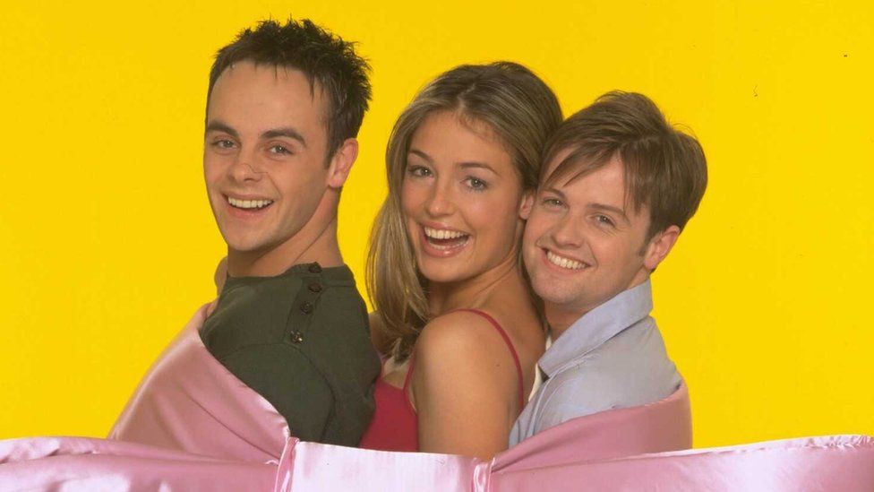 SMTV Live presenters Cat Deeley, Declan Donnelly and Anthony McPartlin, circa 2000. The trio are pictured tied together with a huge pink bow in front of a bright yellow back drop. Ant, wearing a green top, is at the front with Cat sandwiched between him and Dec who stands at the back, wearing a pale blue shirt, his hair cut long over his face. Cat, who has long blonde hair, wears a red strappy top.