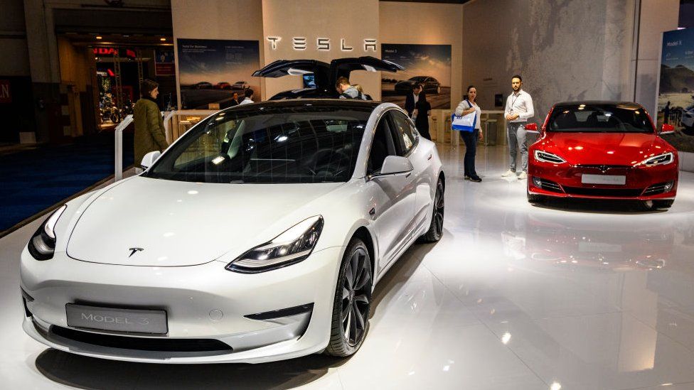 A white Tesla model 3 is seen on the left and a Red Model S on the right on a show floor