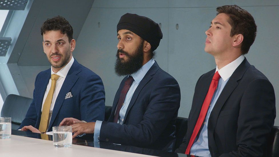 Virdi, an Asian man wearing a dark blue suit and maroon tie, and black turban, sitting in the middle of a boardroom between contestants Steve Darken and Oliver Medforth who are also wearing dark