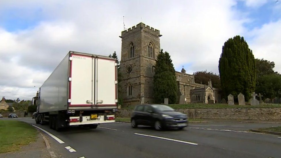 Lorry and car pass on road in the foreground. Isham church tower in the background.