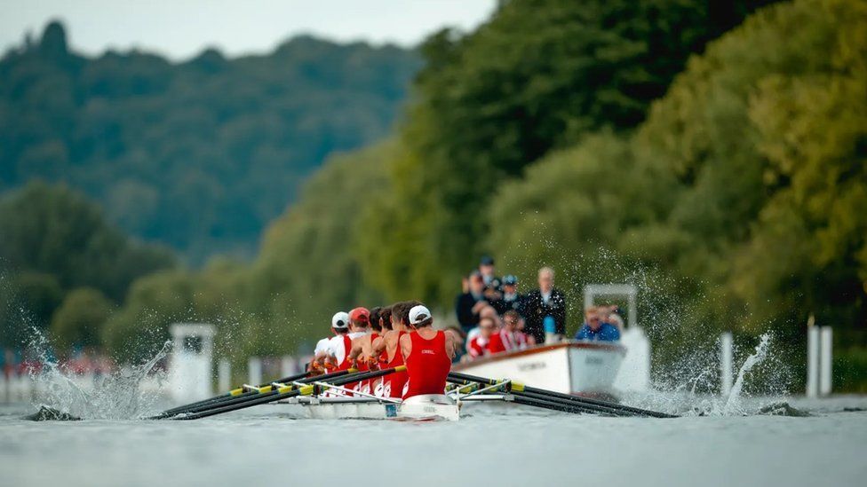 Backs of rowers wearing red race suits with oars splashing and white umpire boat in the background