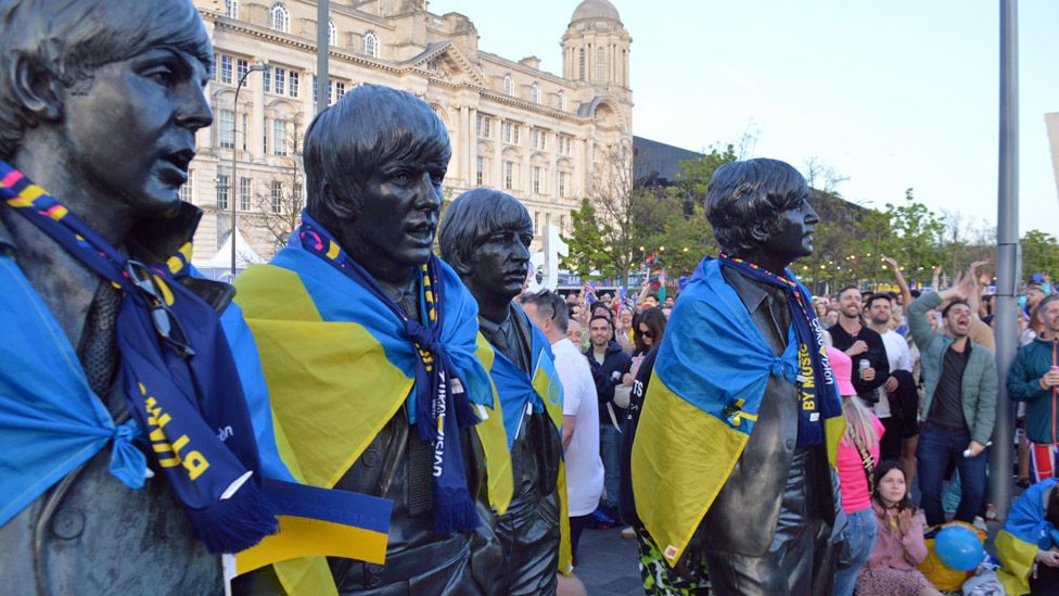 Beatles statues in Liverpool with Ukraine flags and Eurovision scarves around their shoulders, in front of fans in the fan village