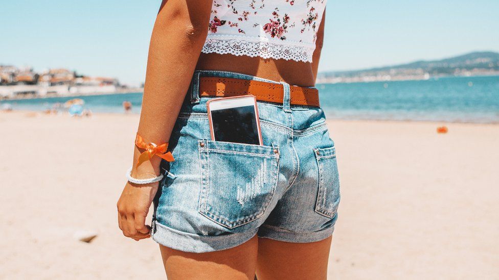 A woman on a beach with a phone in the back pocket of her denim shorts.