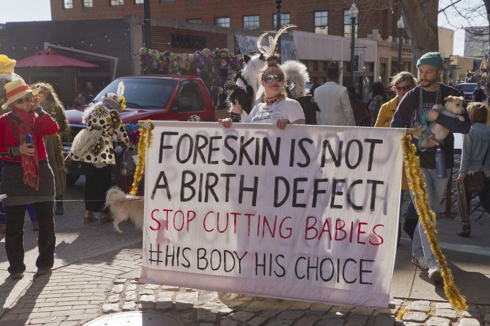Costumed young woman holds large sign about circumcision saying 'Foreskin is not a birth defect' in the Mardi Gras parade on February 7, 2016 in downtown Asheville, NC