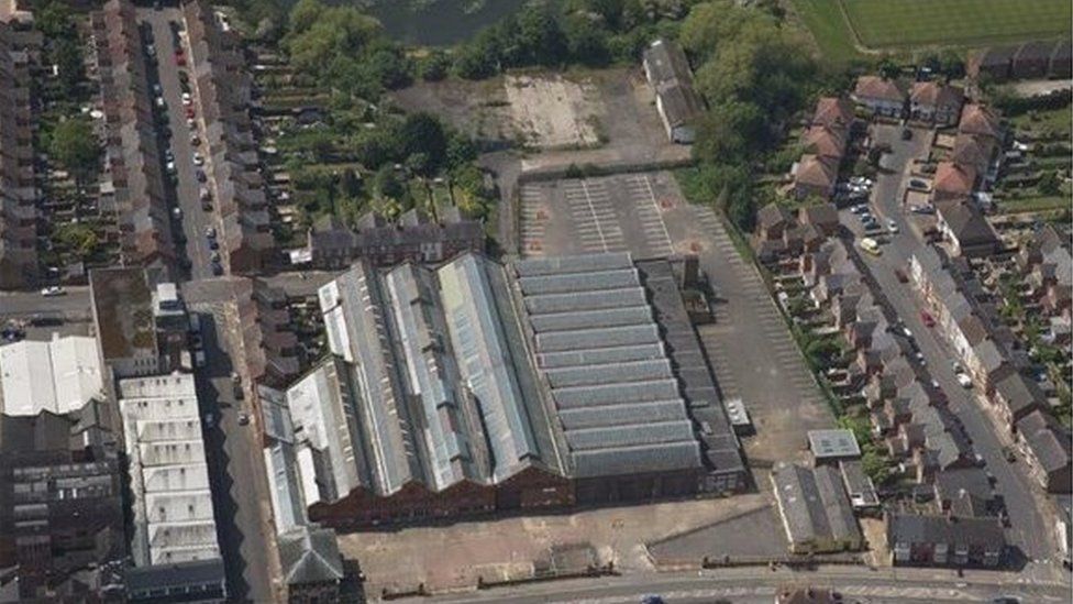 Aerial view showing a line of bus garages with grey roofs, surrounded by houses and trees