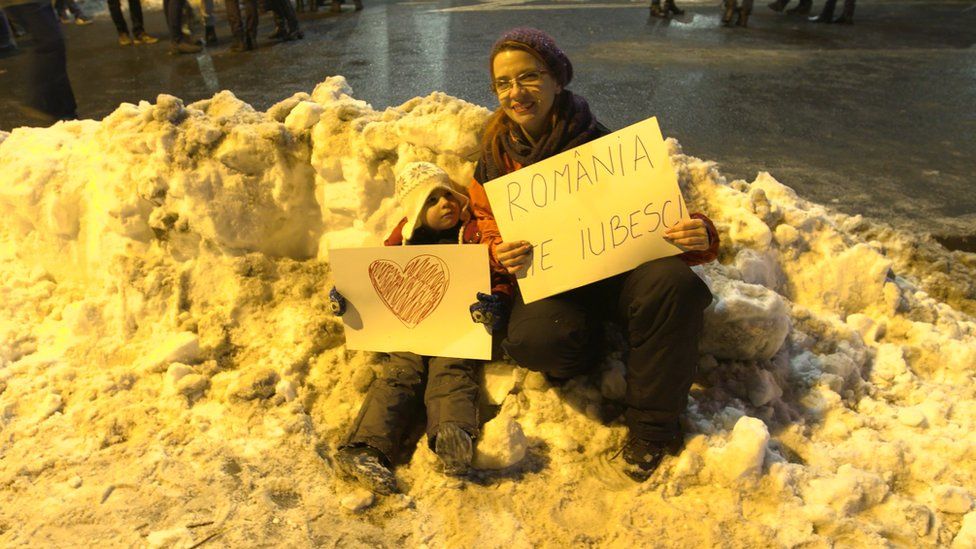 A mother and her young child carrying signs. One reads: "Romania, I love you"