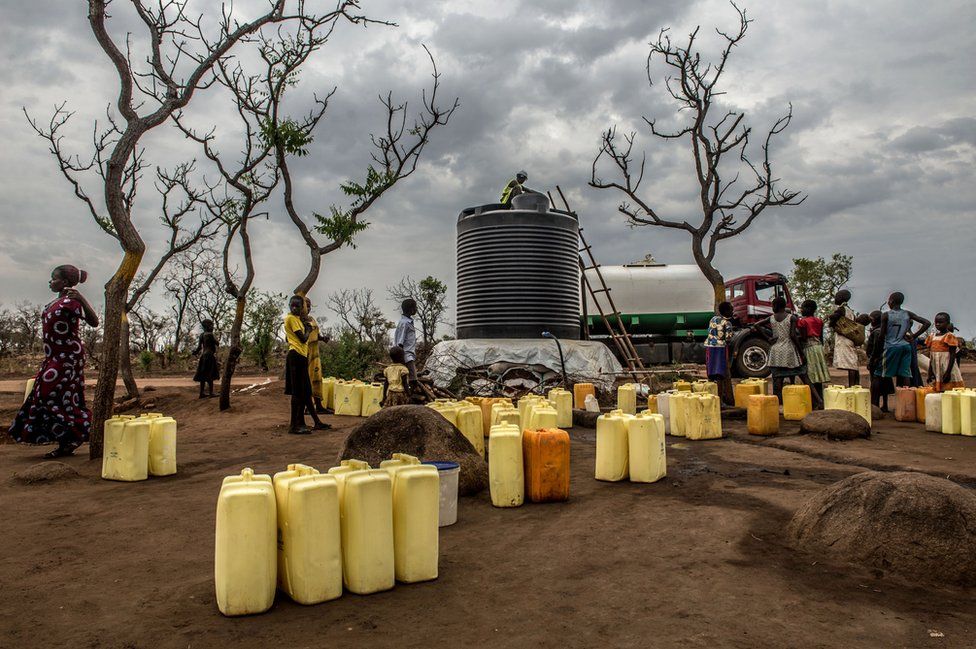 Residents of Rhino camp wait by a water point with the jerry cans, as a tanker offloads its cargo