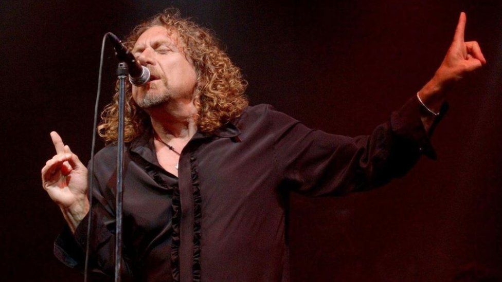Robert Plant singing on stage in front of a mic. Long curly hair, beard, wearing a necklace and a black shirt.