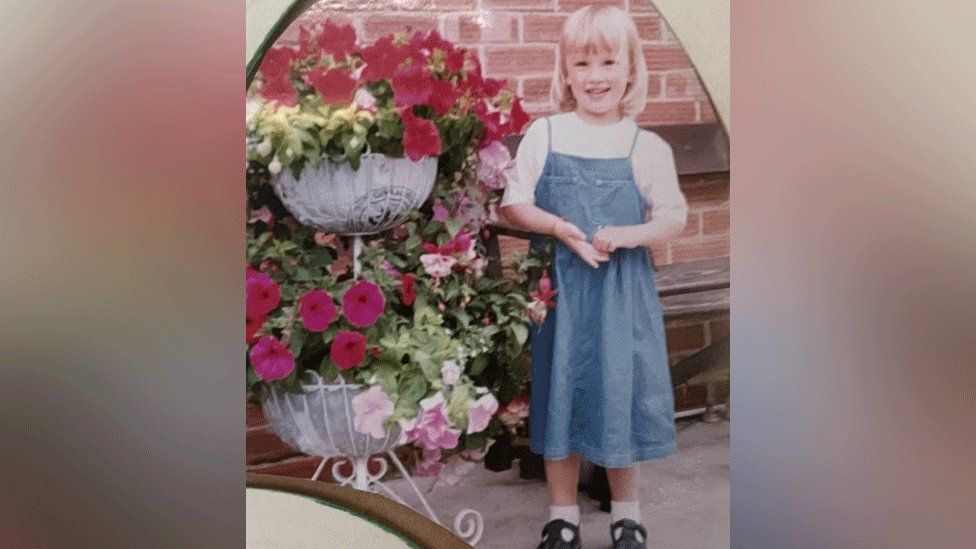 Emily Hales as a child standing next to baskets of flowers on a patio