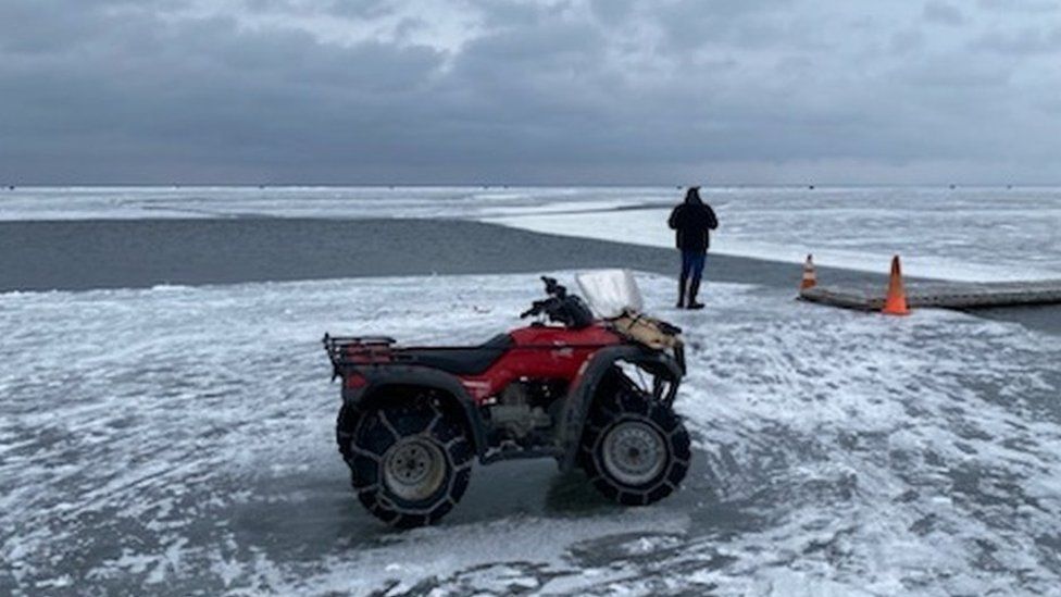 Photo of a rescue vehicle on the ice next to a narrow part of open water with a portable bridge placed atop it so people can cross.
