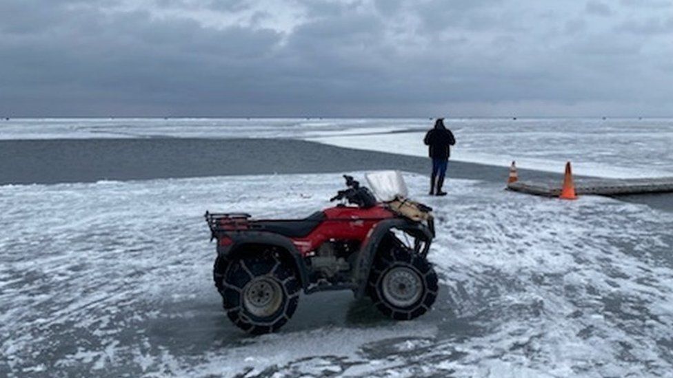 Фотография спасательной машины на льду рядом с узким участком открытой воды с переносным мостом, установленным на нем, чтобы люди могли перейти.