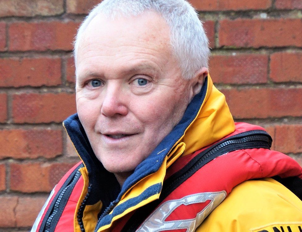 A man in a yellow RNLI jacket and red life preserver standing in front of a brick wall