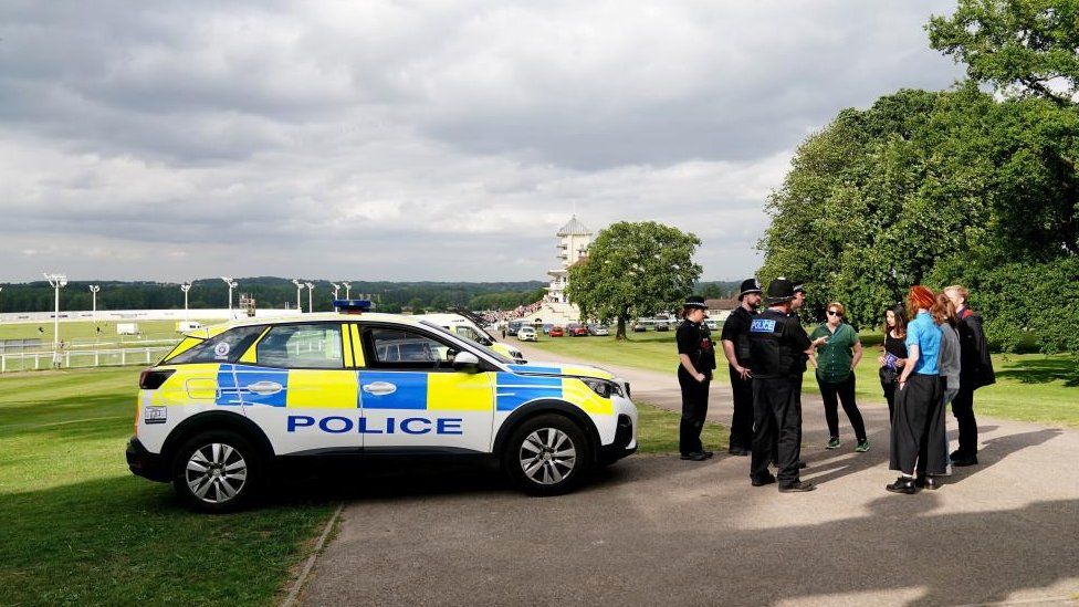 Police officers stand next to a police car talking to a group of people at a racecourse
