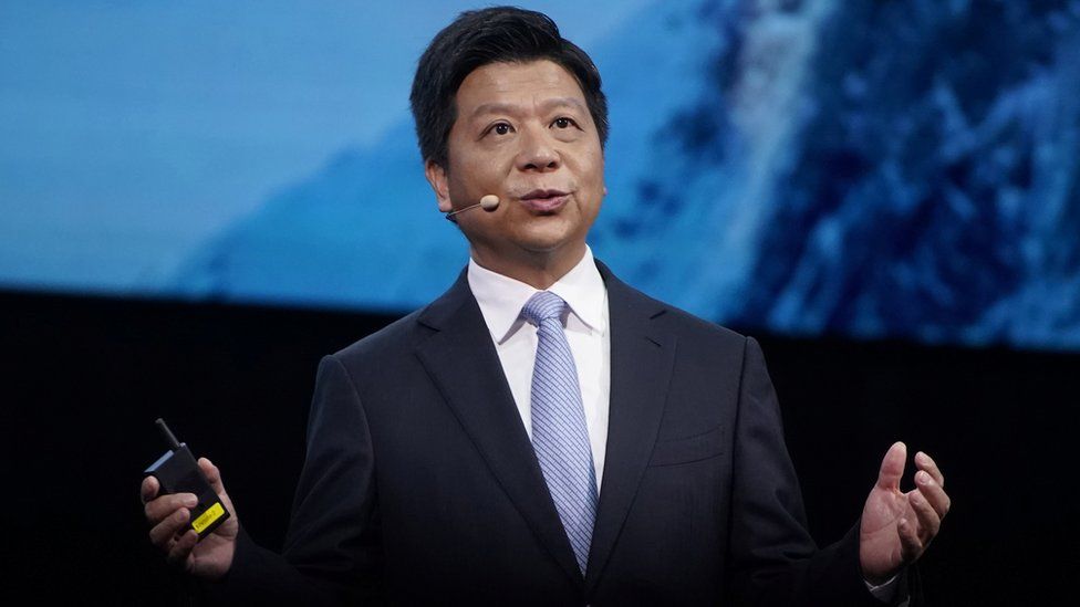 Guo Ping stands in front of a diffused blue photo on a conference stage, arms outstretched while speaking