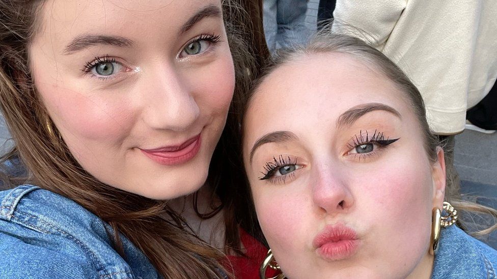 Rosalie Streenstra, left, and her friend at The Weeknd's concert in Amsterdam. Rosalie is a young white woman with blue-green eyes and long brunette hair. She wears a light make up including a glittery silver eye shadow in her inner eye. Her friend, who has blonde hair tied back and grey-blue eyes, leans into her and pouts at the camera. Both of the girls are wearing denim jackets and are pictured in a crowd in the daylight