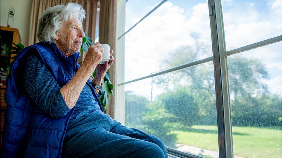 Elderly lady looks out window of care home while drinking a cup of tea
