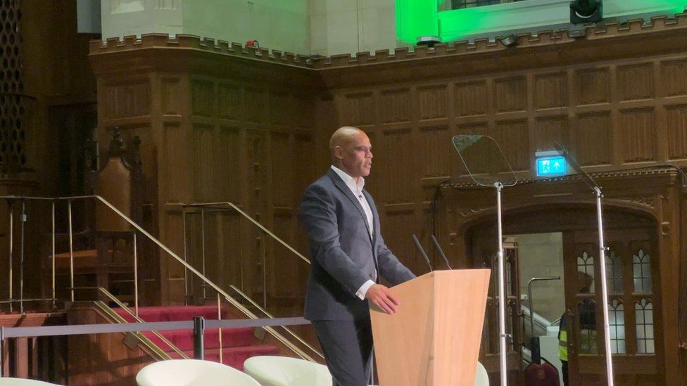 Marvin Rees with a bald head standing at a podium wearing a dark suit and a white shirt addressing a hall