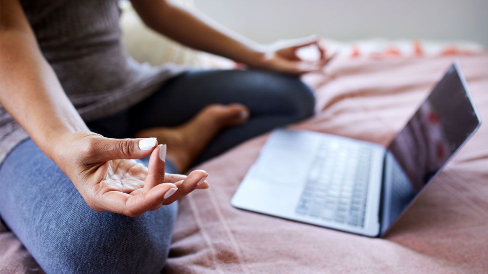 Крупный план женщины, сидящей со скрещенными ногами на своей кровати дома и использующей потоковый класс на ноутбуке, чтобы помочь ей медитировать