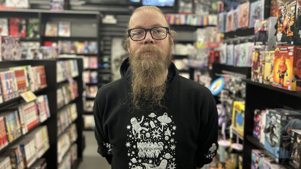 Nick Layland. Nick is a white man in his 30s with long fair hair tied back and a long beard down to his chest. He has blue eyes framed by black rimmed glasses and wears a black hoodie with a cartoon space scene and 'worlds apart' printed on it. He's pictured in a comic book shop with books and figures for sale around him.