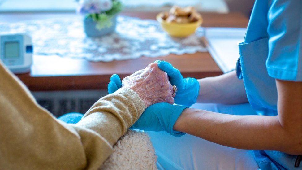 Carer wearing PPE and holding hands with woman in care home