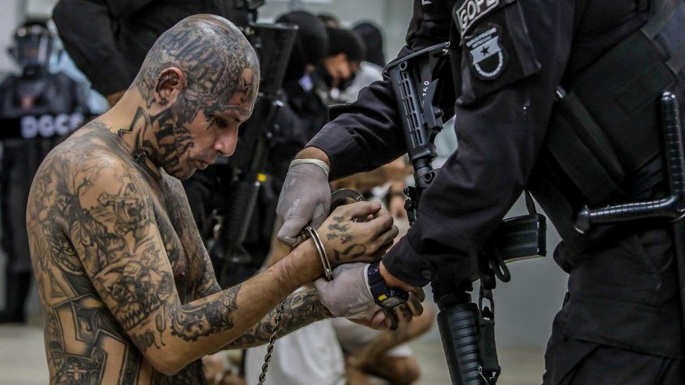 Un agente penitenciario protege a un pandillero mientras lo procesan a su llegada después de que 2000 pandilleros fueran trasladados al Centro de Confinamiento contra el Terrorismo,