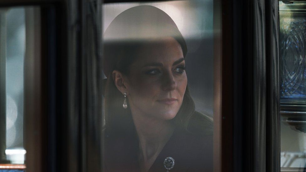 The Princess of Wales, Kate Middleton, looks out of a carriage window. She is wearing a hat, red coat and a brooch with diamonds. She also has pearl earrings on.