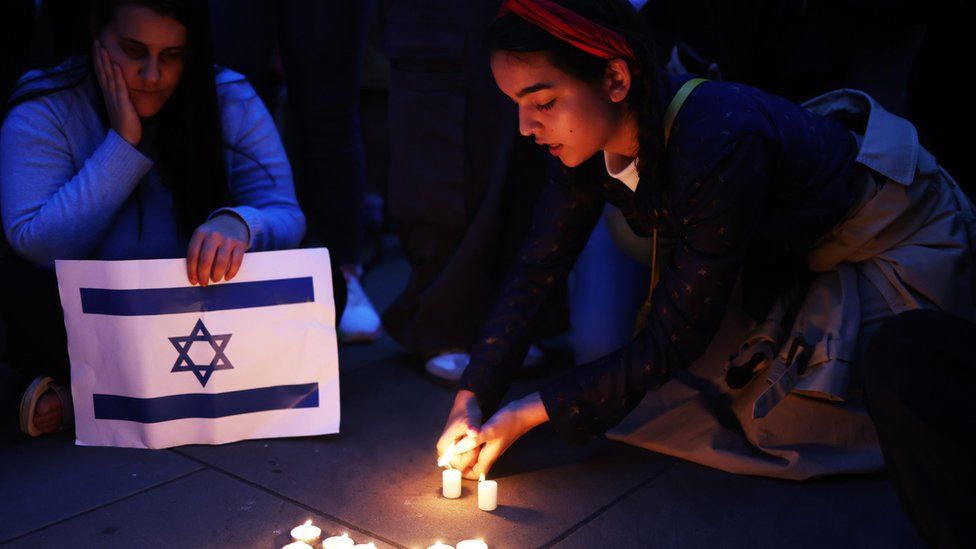 A woman lights a candle while another holds an Israel flag as part of the vigil