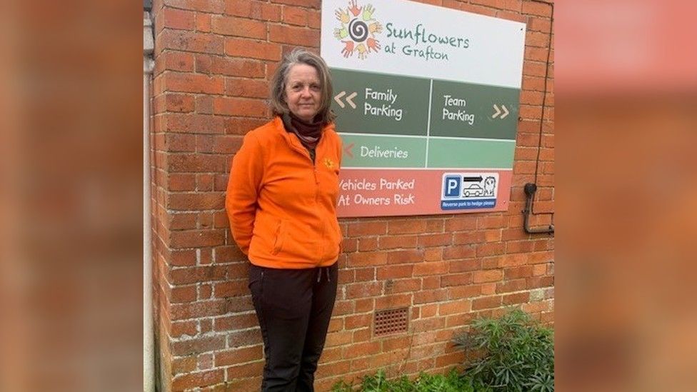 Alison Halfpenny, wearing an orange fleece, standing in front of the nursery's sign on a brick wall