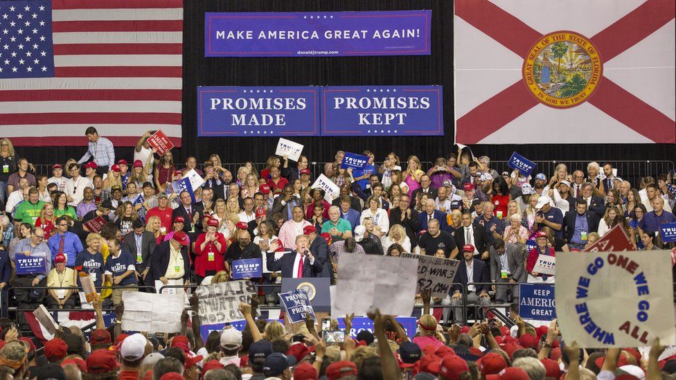 As President Trump speaks at a rally in Florida on 31 July, a QAnon poster is visible (bottom right)
