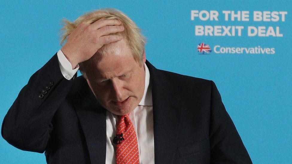 Boris Johnson puts his hand on his head in front of a sign saying "For the best Brexit deal"