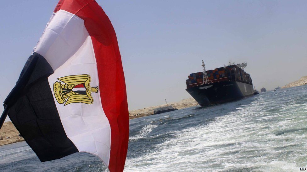 An Egyptian flag flying on the Suez Canal with a cargo ship in the background