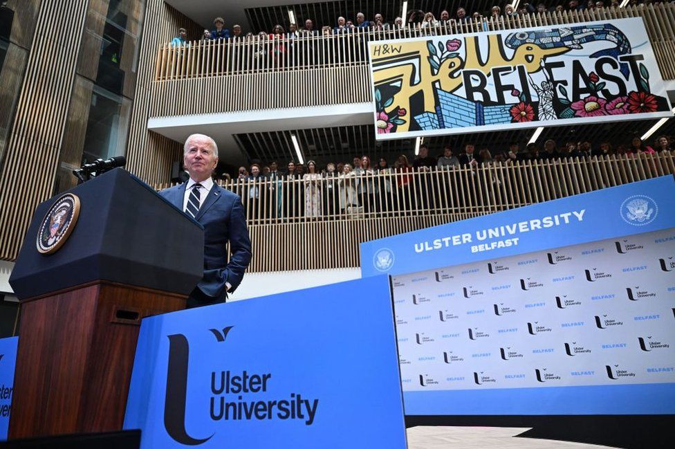 Joe Biden stands at a lectern as he gives a speech at Ulster University in Belfast while dozens of people watch