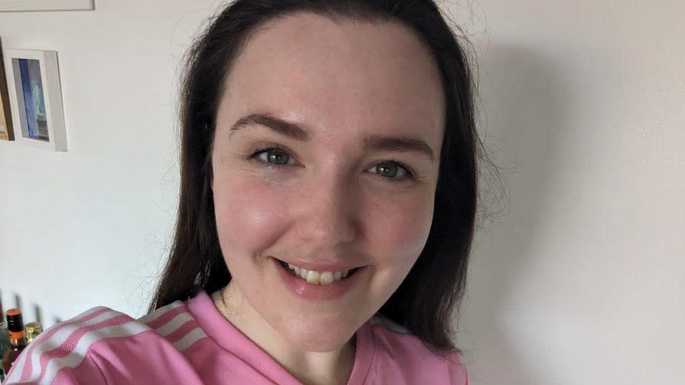 Emma Bowell smiling for a selfie, with a white wall behind her. She is a white woman with long dark hair and is wearing a pink top with three white stripes on the shoulders.