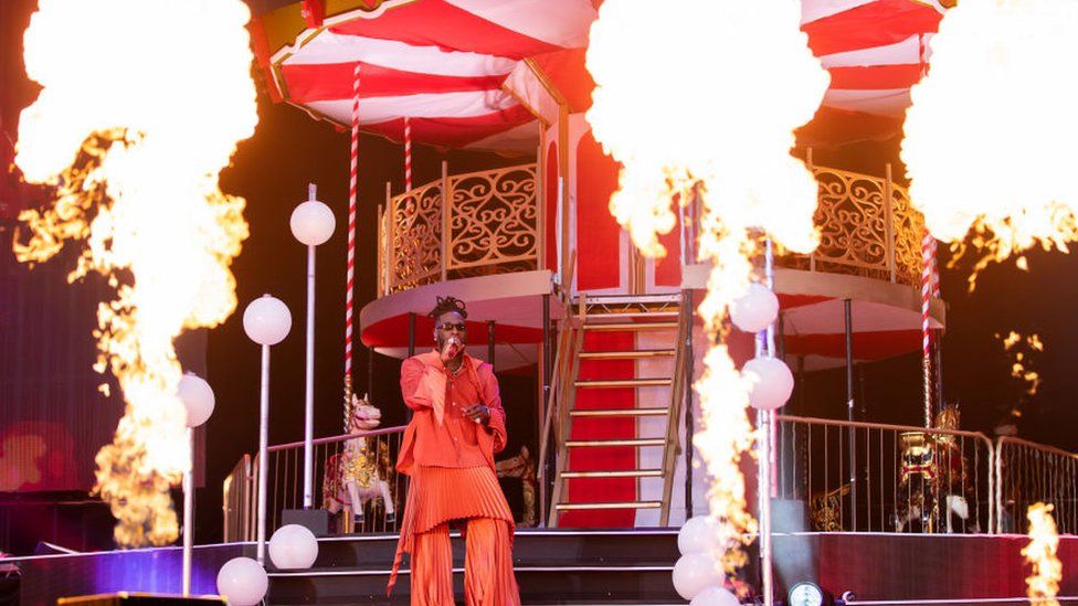 Burna Boy on stage at the London Stadium. He's wearing an orange jumpsuit and behind him on the stage is a fairground carousel and three big lots of flames shooting into the air