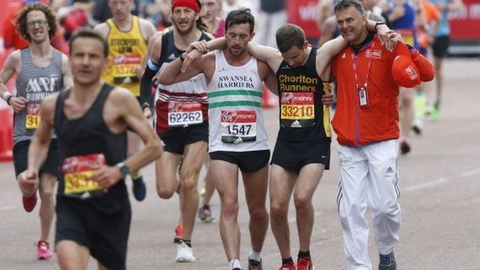 Matt Rees and a race volunteer assist David Wyeth over the finishing line of the London Marathon in 2017