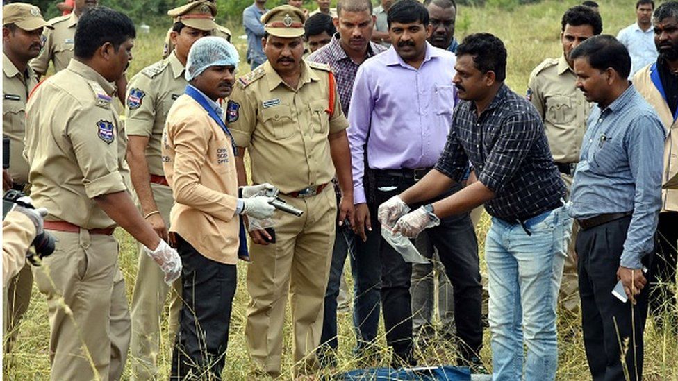 Судебно-медицинский эксперт ((3L) держит пистолет, когда полицейские собираются вокруг тела мужчины на месте, где полицейские стреляли погибли четверо подозреваемых в групповом изнасиловании и убийстве в Шаднагаре, примерно в 55 километрах от Хайдарабада, 6 декабря 2019 г.