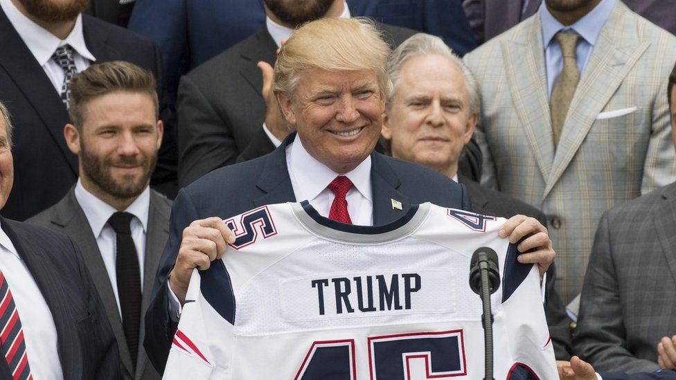 Mr Trump, smiling, holds a "Trump" NFL jersey with the number 45, standing on a podium in front of assembled New England patriots players and staff