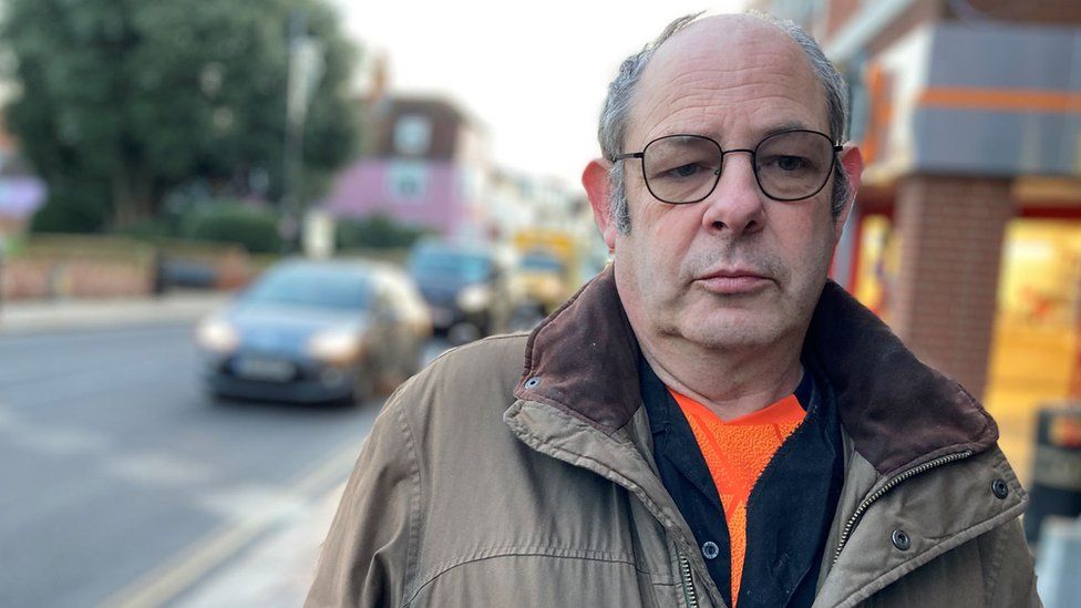 A white man looking at the camera. He is wearing glasses with a black frame a coat and an orange shirt.